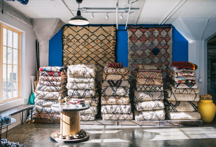 Kechmara Rugs stacked in showroom with large rugs on wall in background
