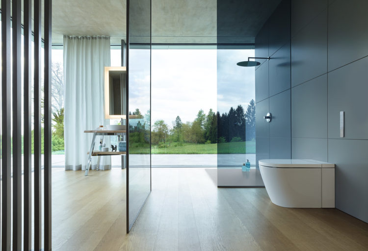 Duravit SensoWash i side view of toilet in open bathroom with glass-walled shower and exterior green hill and trees visible out window