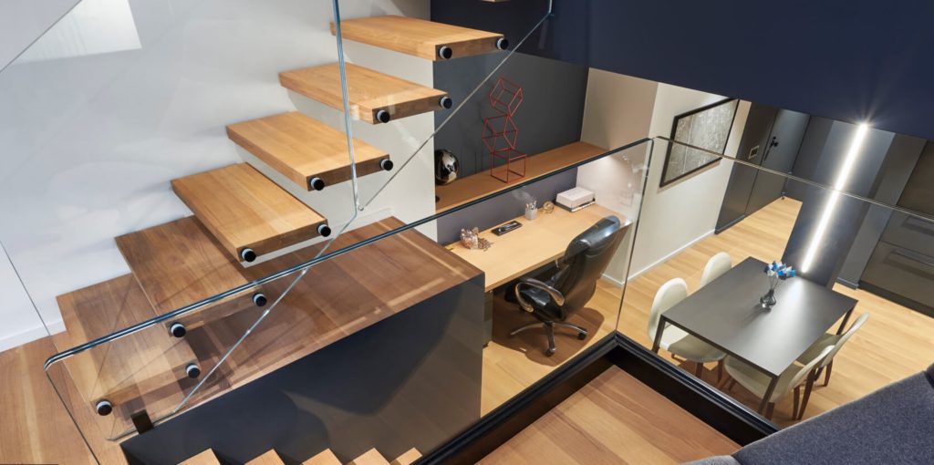 Staircase with broad wooden treads and glass panel style railing in small apartment.