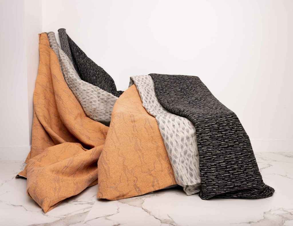 NeoCon Wolf-gordon Muse Collection textiles three styles orange/yellow with Eqyptian woman motif, cream/gray with curvy shapes, and black with brown horizontal lines