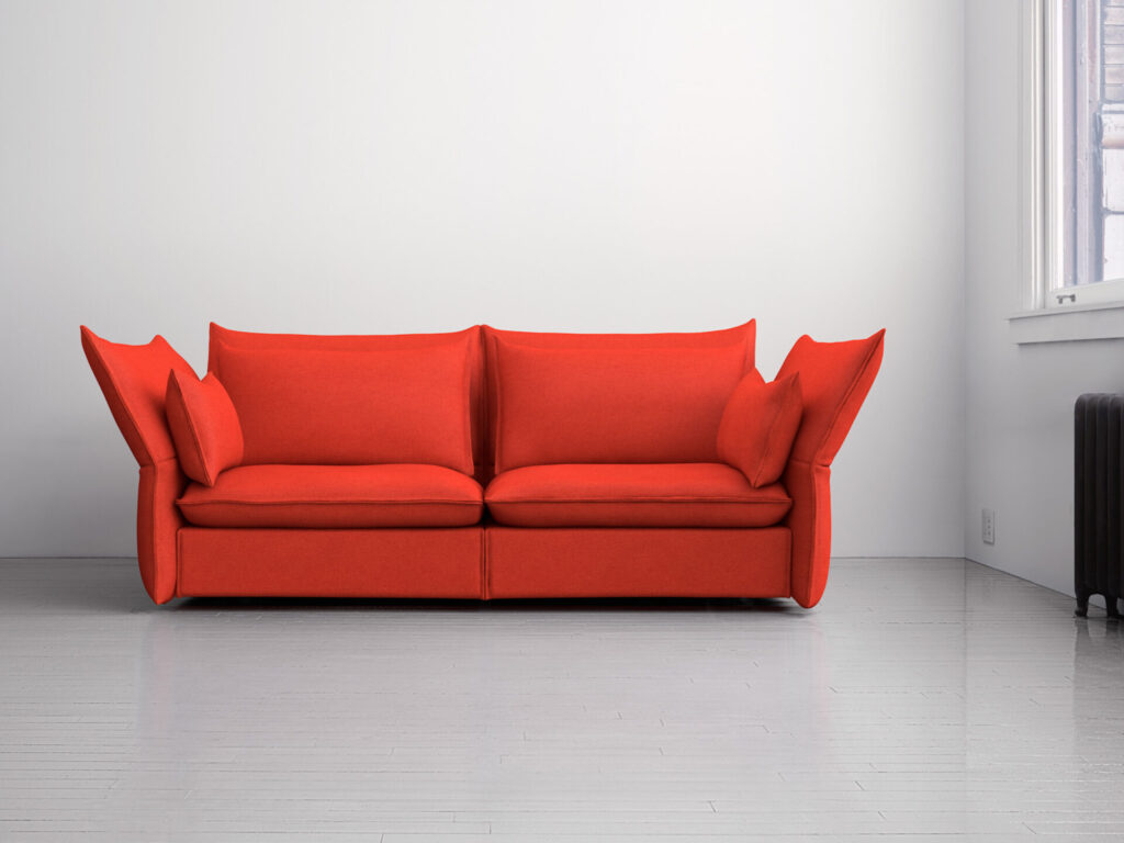 Modern three-seat sofa upholstered in bright red fabric, featuring oversized back and side cushions with a sculptural, folded appearance. The sofa sits on a light gray wood floor in a minimal white room with natural light coming from a window on the right. Upholstery shown is Maharam’s Messenger.