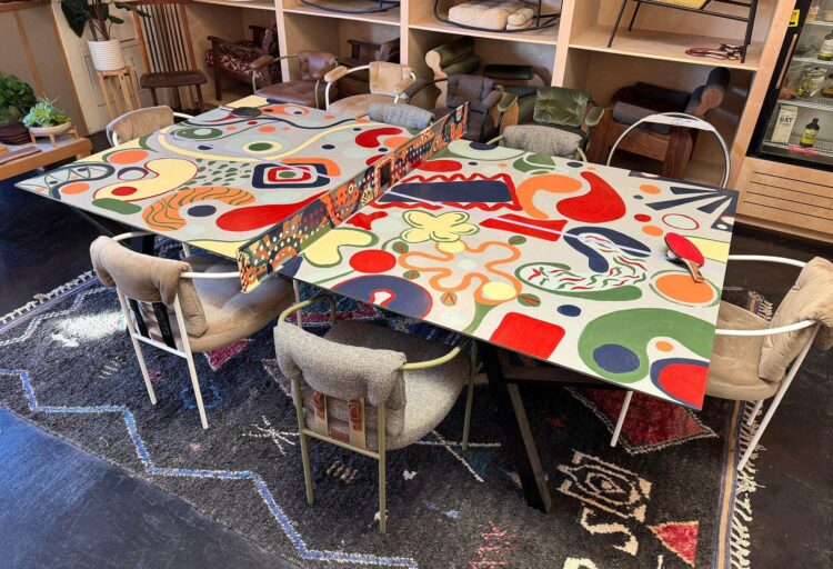 Detail view of the table’s hand-painted steel net. Circular perforations allow visibility through the sculptural surface, while the Sean Woolsey Studio logo medallion anchors the vibrant palette.