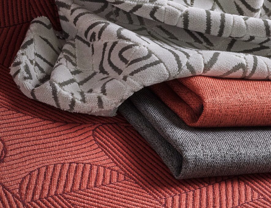Stack of neatly folded upholstery textiles in various colors and woven patterns, including geometric and organic motifs. The fabrics are part of Stinson’s “The Collective” collection, presented in a soft, layered composition.