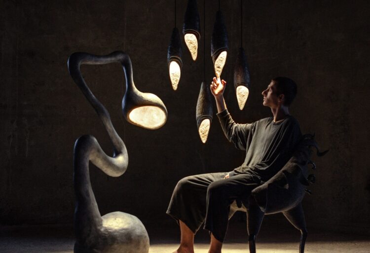 A man sits barefoot in a dimly lit space, surrounded by organic sculptural lamps—one twisting floor lamp and a cluster of cocoon-like pendants—bathed in warm light.