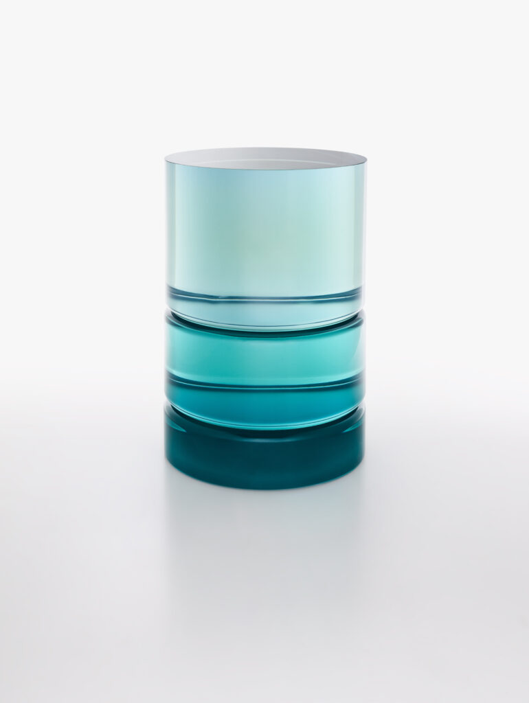 A cylindrical ETS resin side table in teal and aqua tones displays precise banding and gradient transitions, turning volume and translucence into an exercise in architectural color.