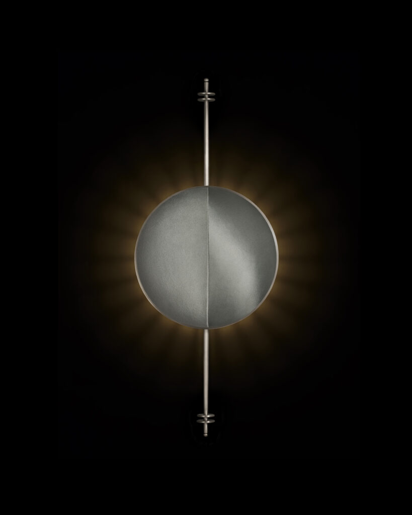 A circular suede-wrapped wall sconce glows like an eclipse, with a halo of warm light radiating behind its muted surface.