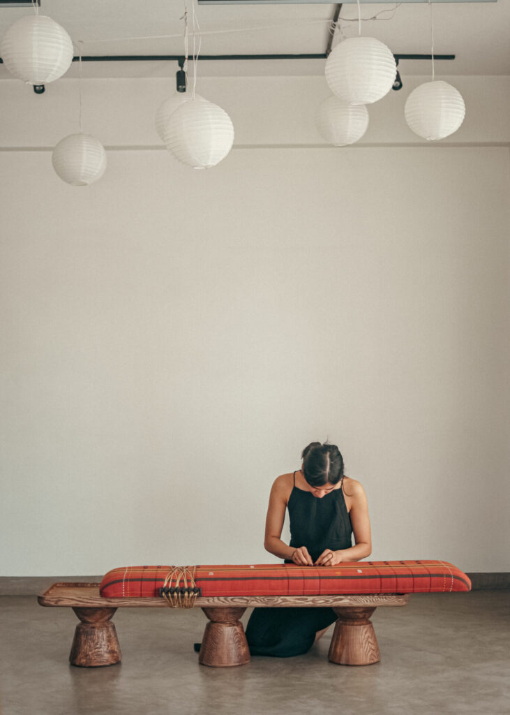 Designer seated at a long wooden bench with handwoven rust-and-mustard cotton seat, lashing cane ties across the frame beneath hanging paper lanterns.