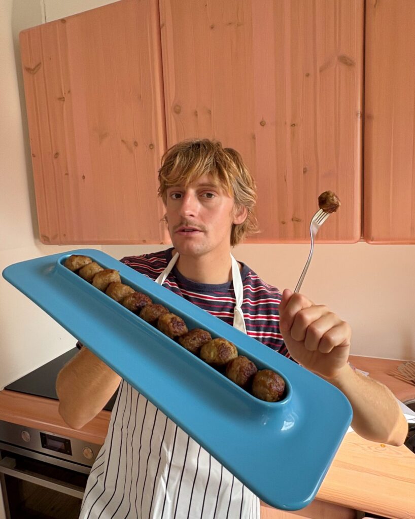 A person in a striped shirt and apron holds the turquoise meatball plate filled with a straight row of cooked meatballs, lifting one on a fork.