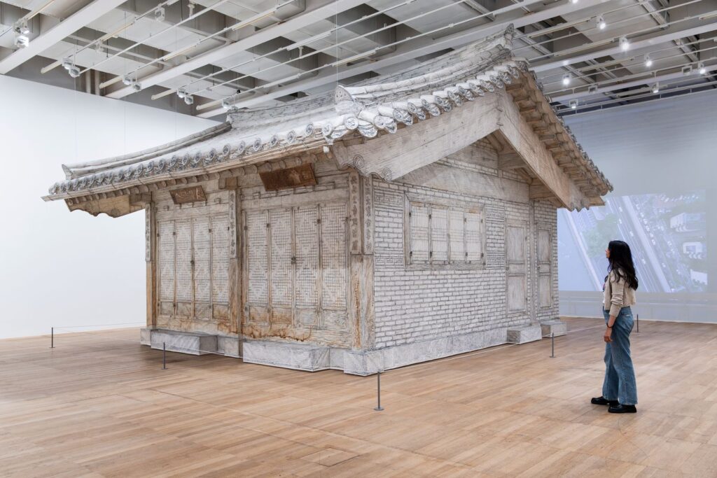 A life-size architectural sculpture of a traditional Korean house, rendered in pale graphite-like textures, fills a gallery as a visitor observes its intricate details.