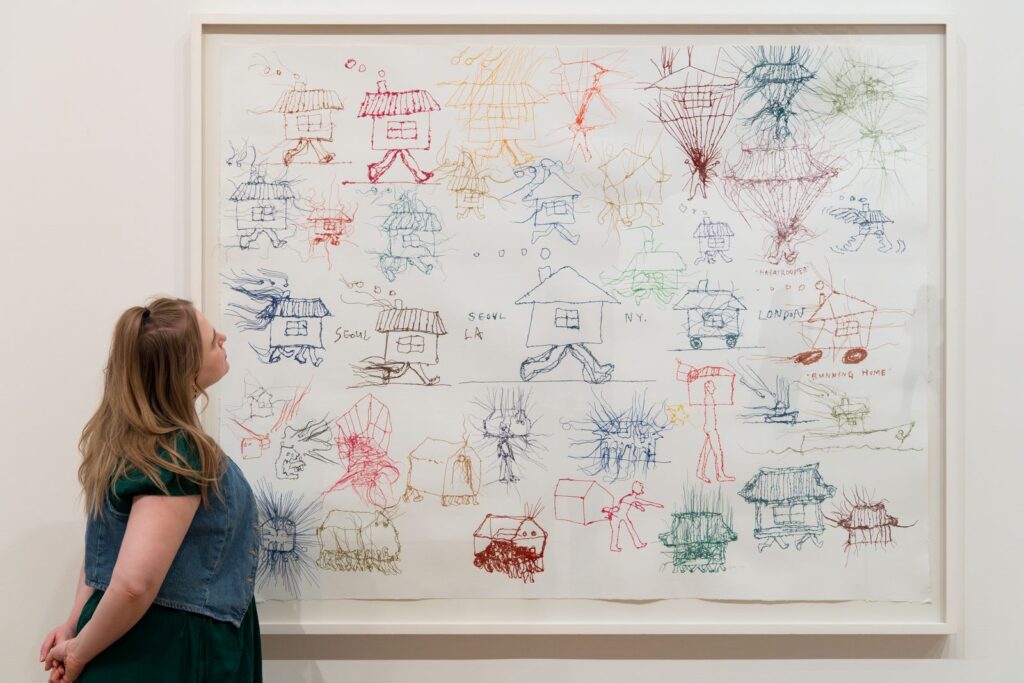 A woman studies a framed artwork filled with colorful thread sketches of walking houses and figures, each labeled with cities like Seoul, London, and New York.