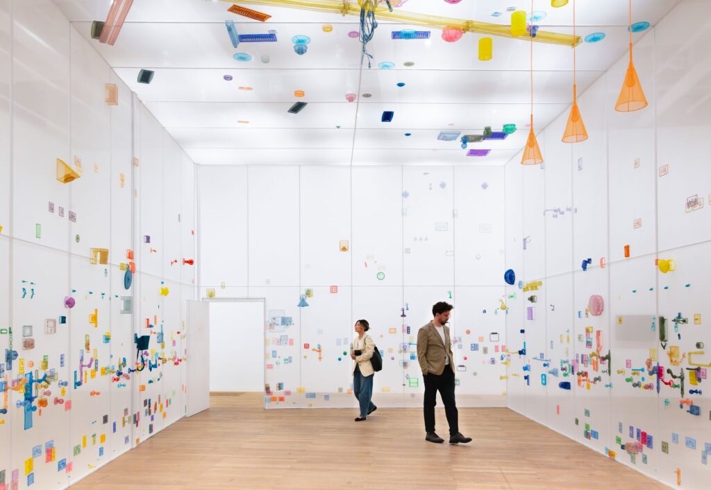 Two visitors walk through a white gallery where translucent plastic household objects in bright colors are suspended on walls and ceiling, creating a playful, immersive installation.