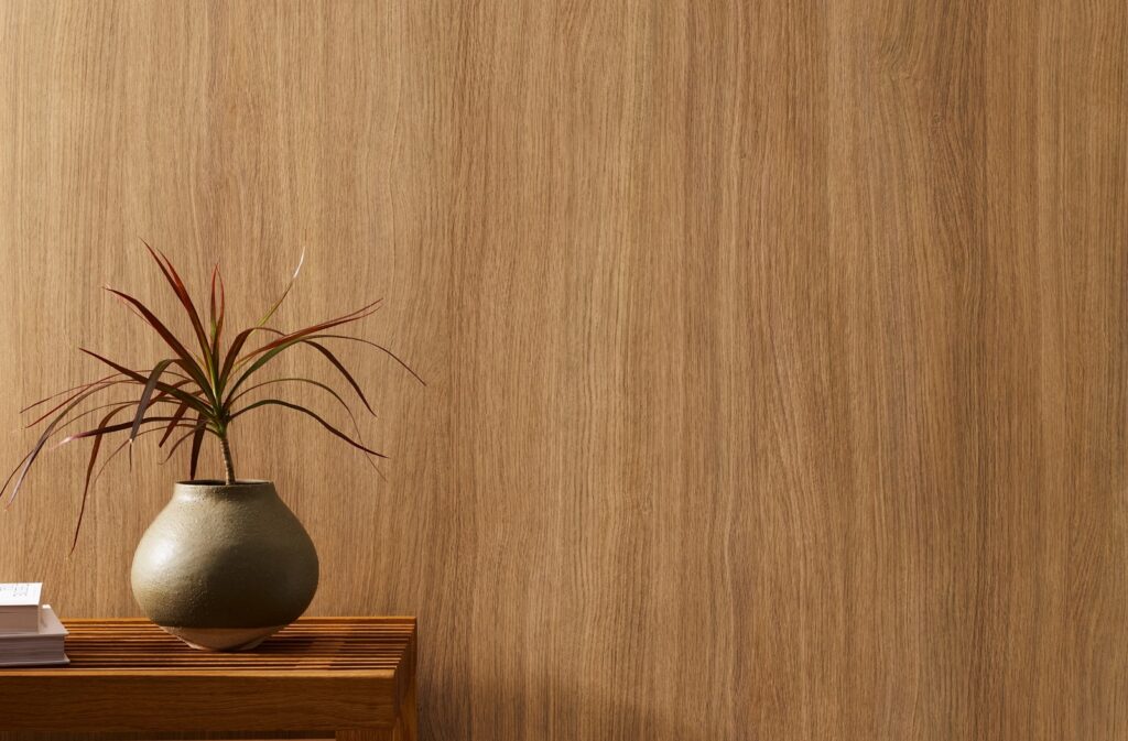 Warm honey-toned wood-grain wallcovering with subtle linear texture, styled with a sculptural ceramic vase and spiky dracaena on a slatted wood bench.
