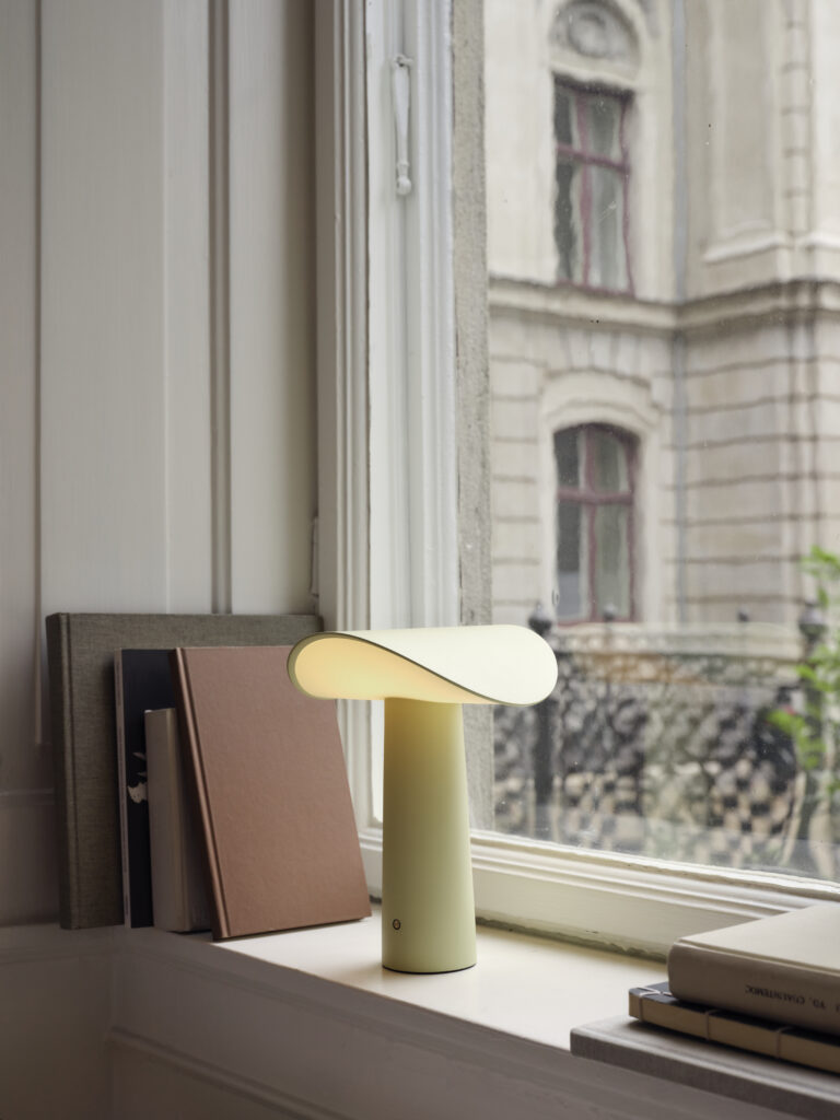 The Lium portable lamp in pale yellow sits on a windowsill beside stacked books, its curved shade casting a warm glow against the urban backdrop.