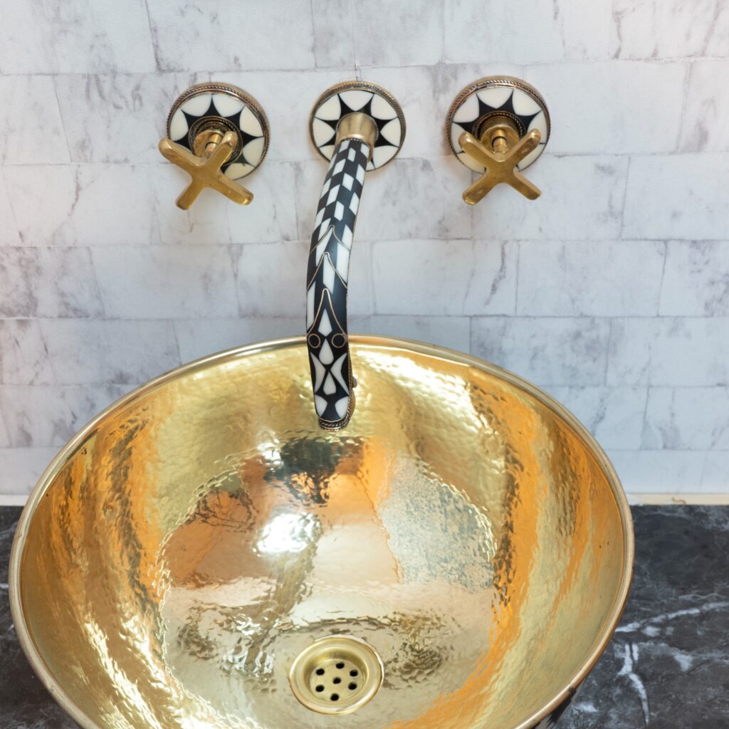 A hammered brass vessel sink glows with molten warmth beneath a wall-mounted faucet, its spout inlaid with black-and-white geometric patterns that turn plumbing into ornament.