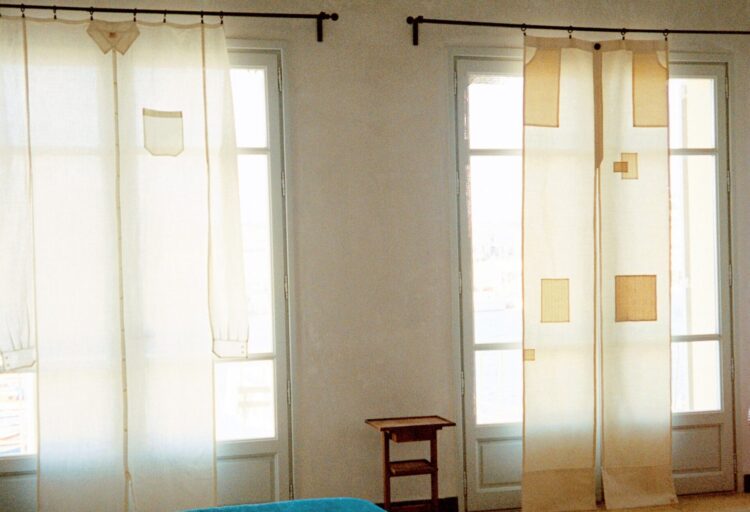Two sheer linen curtains styled with shirt details—collar, cuffs, and a breast pocket—hang across tall windows, turning a bedroom into a playful dressing room for architecture.