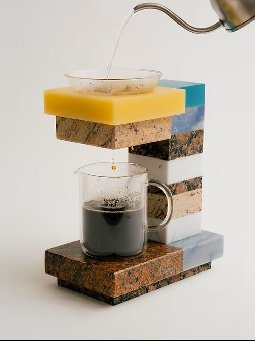 Hot water pours from a gooseneck kettle into the glass dripper of the Stone Drip brewer, coffee collecting below in a clear carafe atop the granite base.