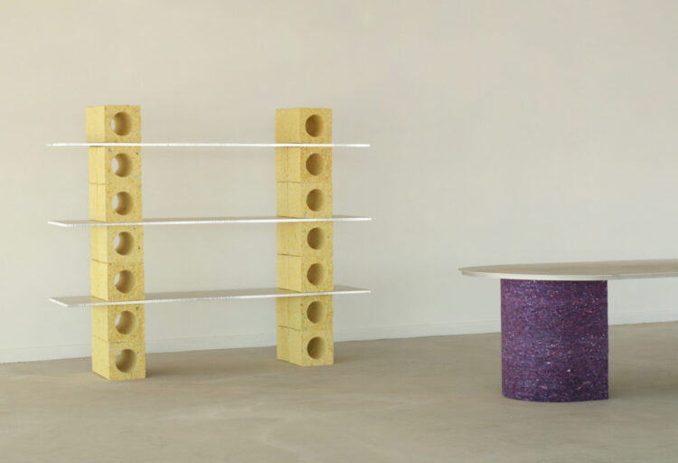 A minimalist studio scene pairs the yellow-block shelving with a long oval table on a deep violet cylindrical base. Together, they form a study in color, geometry, and circular design thinking.