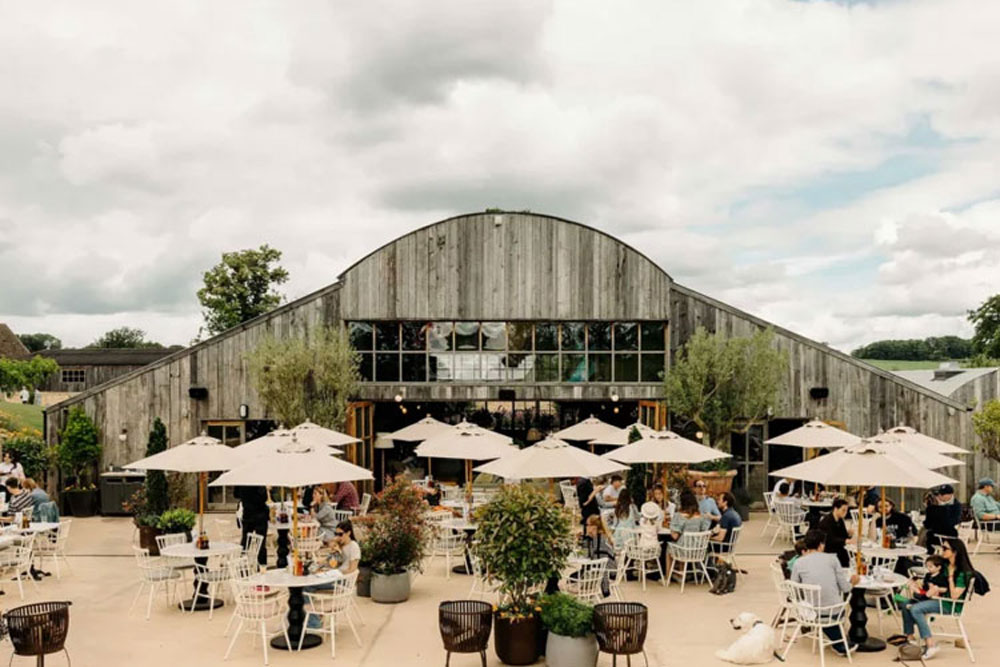 Guests dining on a spacious terrace at Soho Farmhouse, with white umbrellas, outdoor seating, and a rustic barn-style building in the background.