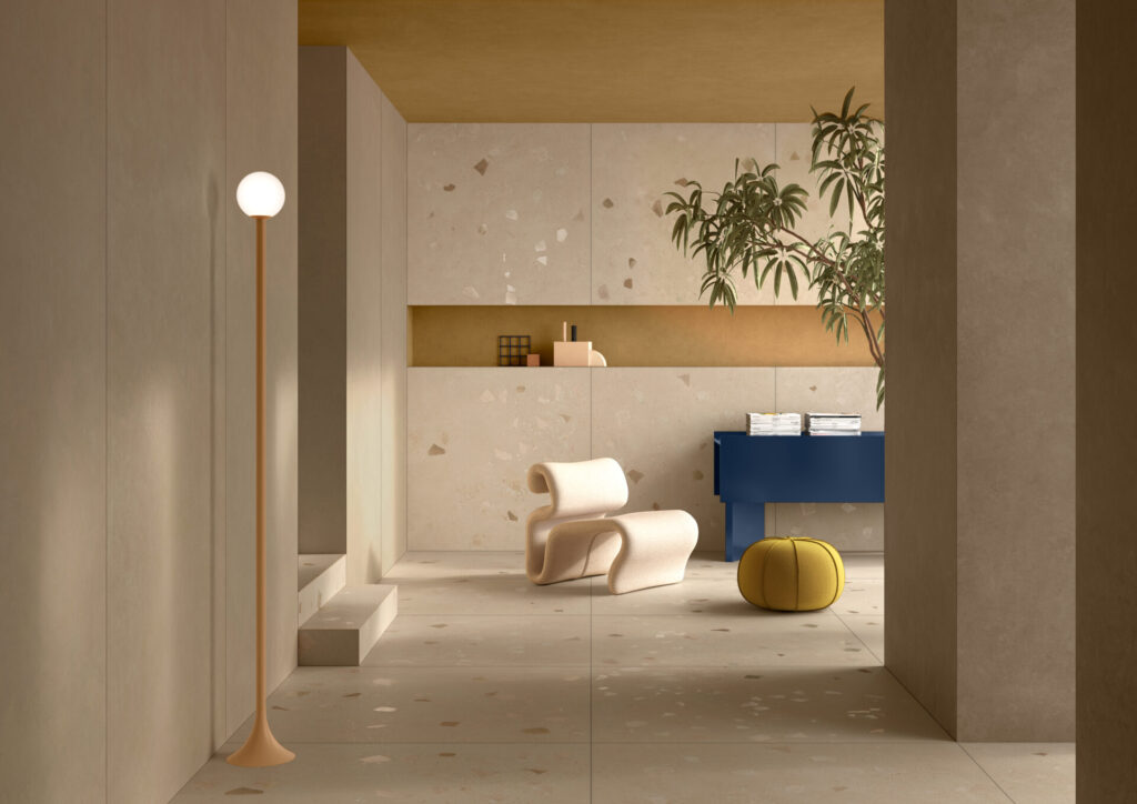 A serene interior hallway finished in pale terrazzo-look tiles with warm undertones. A sculptural cream lounge chair, yellow pouf, and cobalt console bring gentle contrast beneath a mustard ceiling. Light plays across the seamless wall and floor surfaces, underscoring the precision of tone and texture.