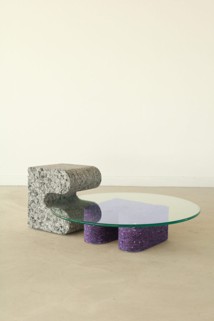 A low coffee table combines a sculpted grey block with a purple recycled-fabric base beneath a round glass top. The piece reads as both monolithic and light, merging raw reuse with refined proportion.