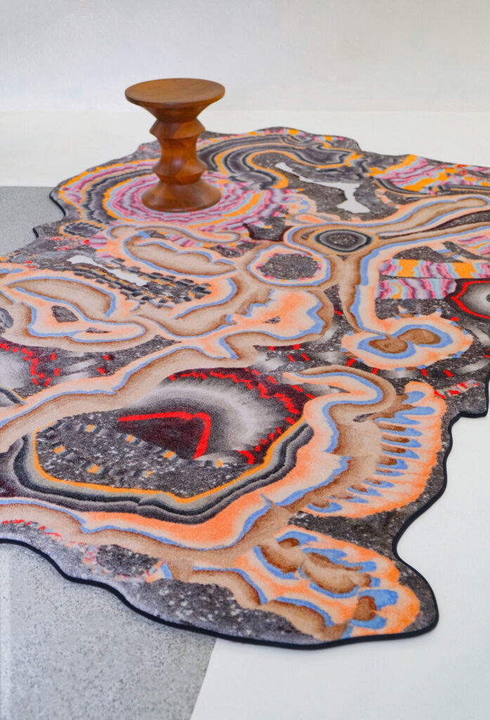 The BAUBAU Fossil Psychic rug by Kerstin Brätsch for ZigZagZurich sprawls organically beneath a sculpted wooden stool, its swirling forms evoking magma and mineral layers in vivid coral and gray.