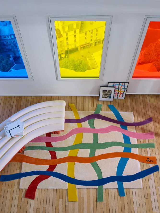 In a sunlit room with blue, yellow, and red tinted windows, the Crossroads rug stretches across wood floors, its multicolored ribbons creating a lively grid beneath a sculptural cream sofa. A framed collage leaning against the wall echoes the rug’s expressive lines.