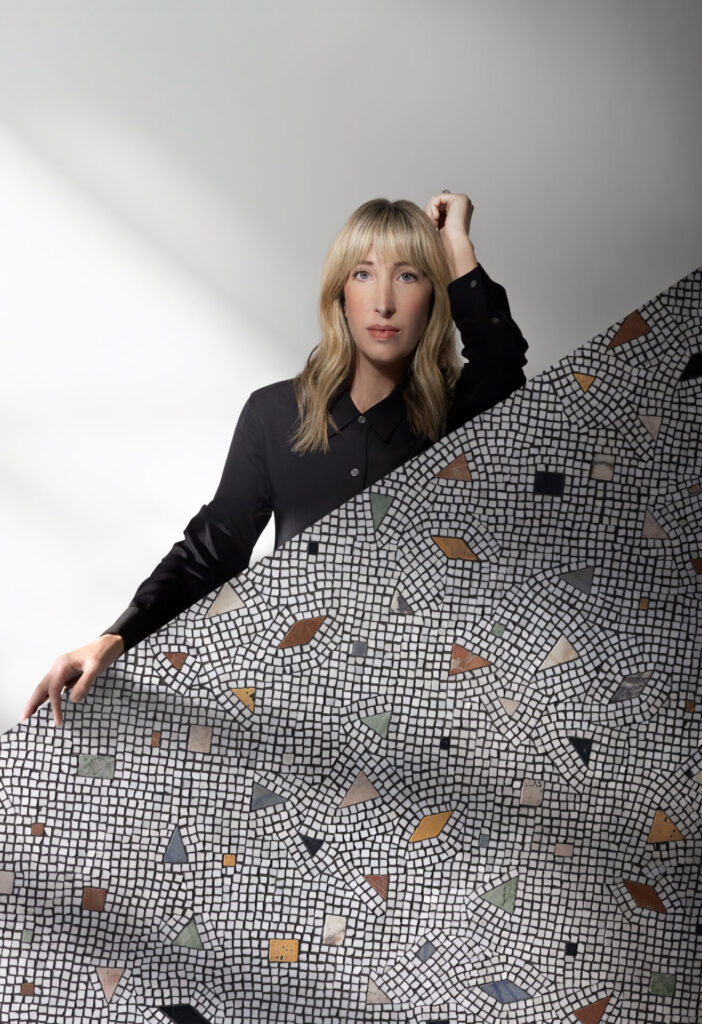 Set against a wash of cool studio light, a designer in a black shirt frames a sheet of Artifact mosaic in deeper neutrals, where tumbled terracotta and sage pieces break through a field of tight black-and-white tesserae. The effect is graphic yet organic, a modern riff on Roman pavement.