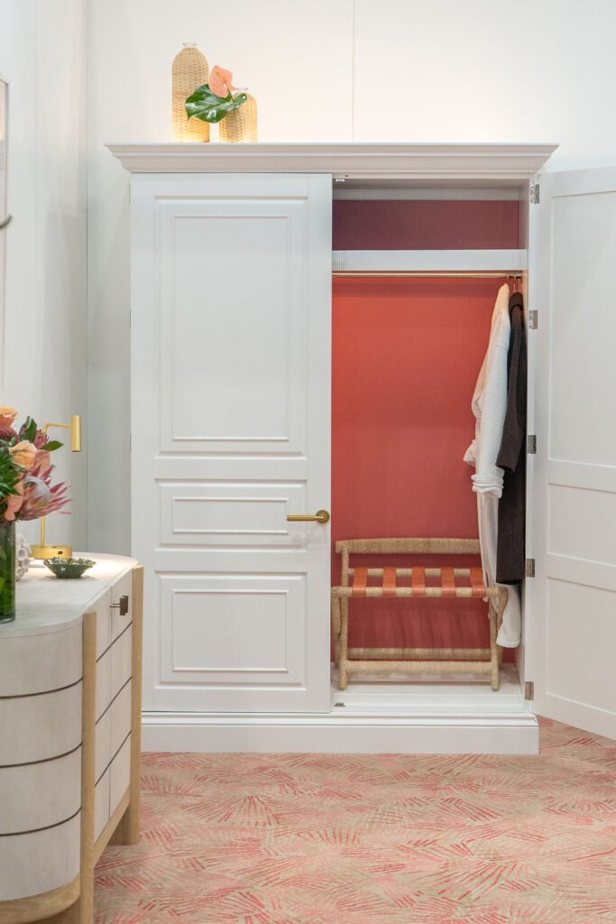 A white paneled wardrobe opens to reveal a coral-washed interior with a woven luggage bench and soft brass hardware, a calm coastal moment inside Bernhardt’s Palm House vignette.