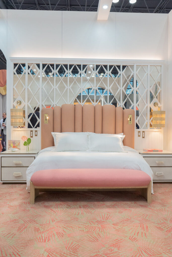 A tall, channel-tufted headboard in sandy peach anchors a serene guest room setting, paired with crisp white bedding, woven bedside lighting, and a pink bench that completes the Palm Beach palette.