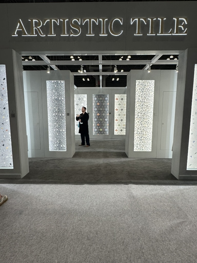 The Artistic Tile booth at BDNY 2025 glows with backlit mosaic panels, each showcasing intricate geometric and organic patterns. A visitor stands centered beneath the illuminated “ARTISTIC TILE” signage, surrounded by towering displays that reveal the brand’s depth in stone craft.