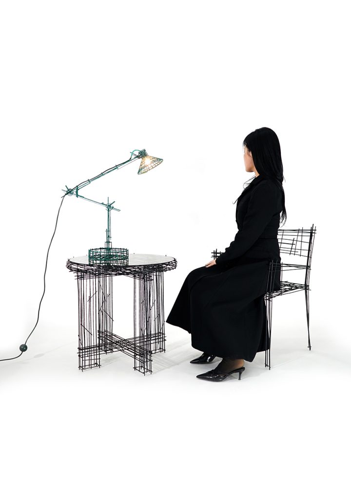 A woman in black sits at a table and chair made entirely of thin, black wire lines, beside a green wire desk lamp—each piece appearing like a sketch come to life.