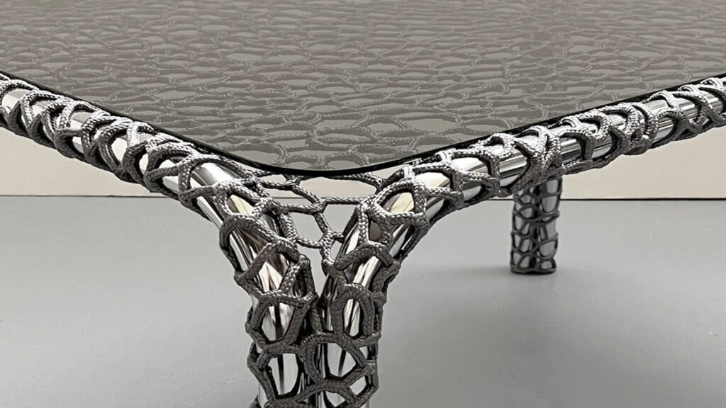 Close-up of a chrome table corner wrapped in hand-spliced charcoal rope, the woven pattern forming an intricate lattice that softens the metal’s industrial precision.