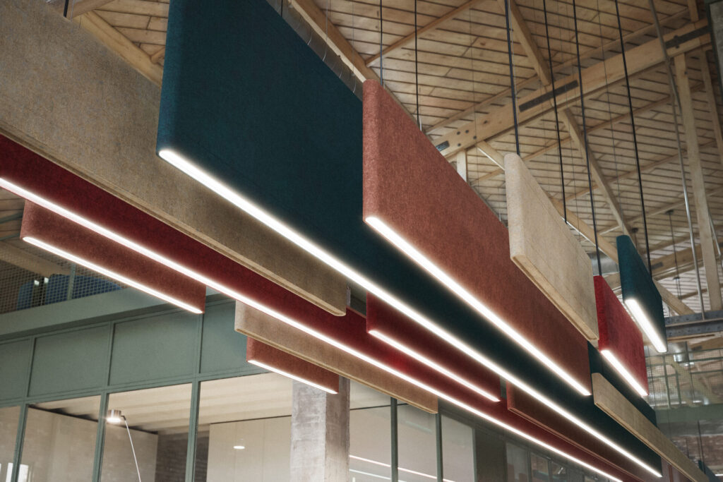 A close-up view of Sabin’s Linear Stadia baffles, layered in deep teal, red, and sand tones. Each rounded felt profile carries a continuous line of integrated lighting, forming a rhythmic overhead canopy that adds warmth and softness to an industrial interior.