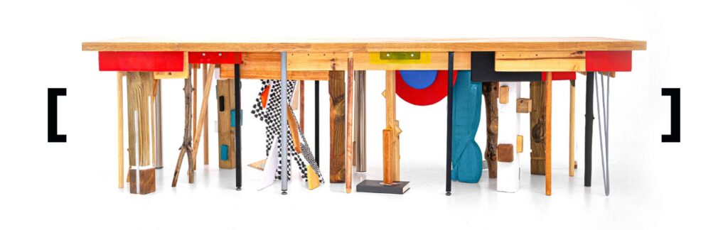 A straight-on view of the same eclectic dining table, its underside packed with vivid, clashing legs in wood, metal, acrylic, and boldly painted forms, turning structural support into a dense, playful collage of materials and shapes.