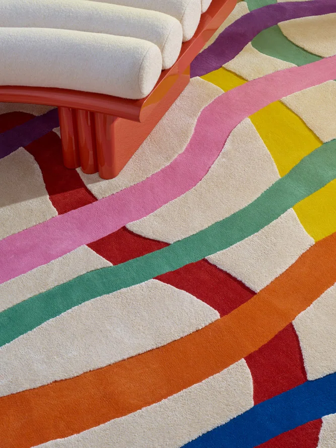 A detailed view of the rug reveals its plush texture and layered color bands—pink, red, green, orange, and blue—intersecting beneath the curved legs of a cream upholstered bench. The fibers catch the light, giving the palette extra depth.
