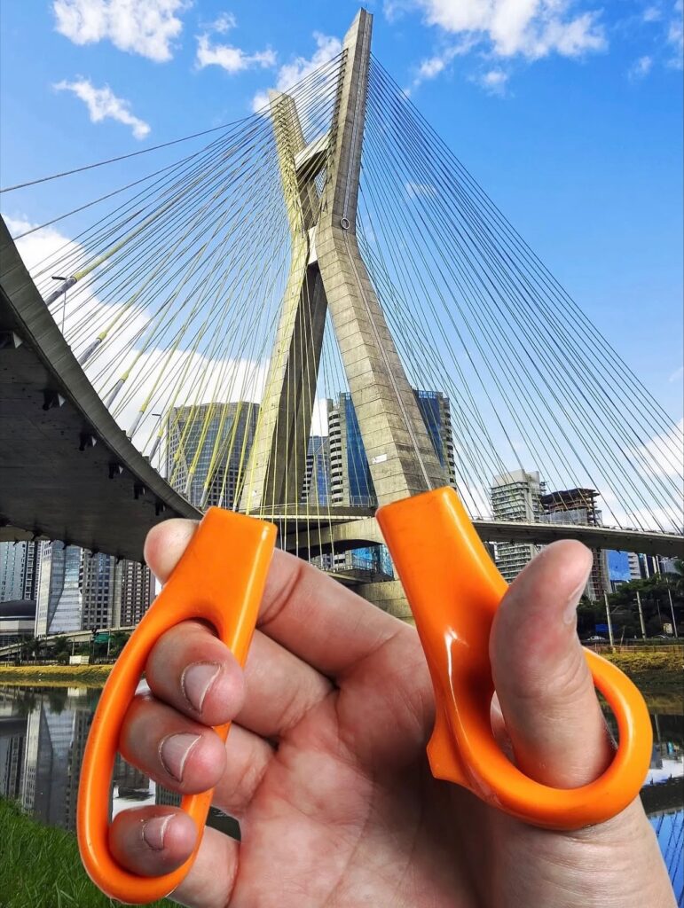 Bright orange scissor handles are held up to São Paulo’s Octavio Frias de Oliveira Bridge, matching the bridge’s split tower so precisely they appear to be its oversized grips. The suspension cables radiate like taut threads ready to be cut.