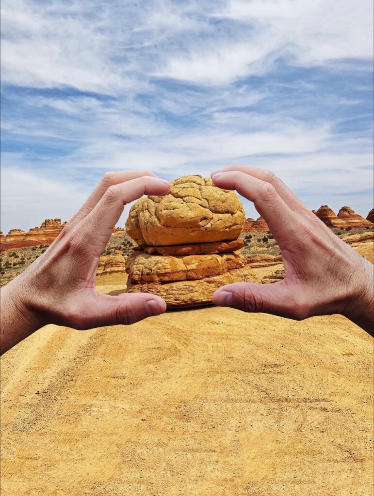 Two hands frame a desert rock formation, the rounded boulder stacked on sandstone layers resembling a giant pastry being gently shaped. The gesture turns geology into something almost edible, a playful take on scale and texture