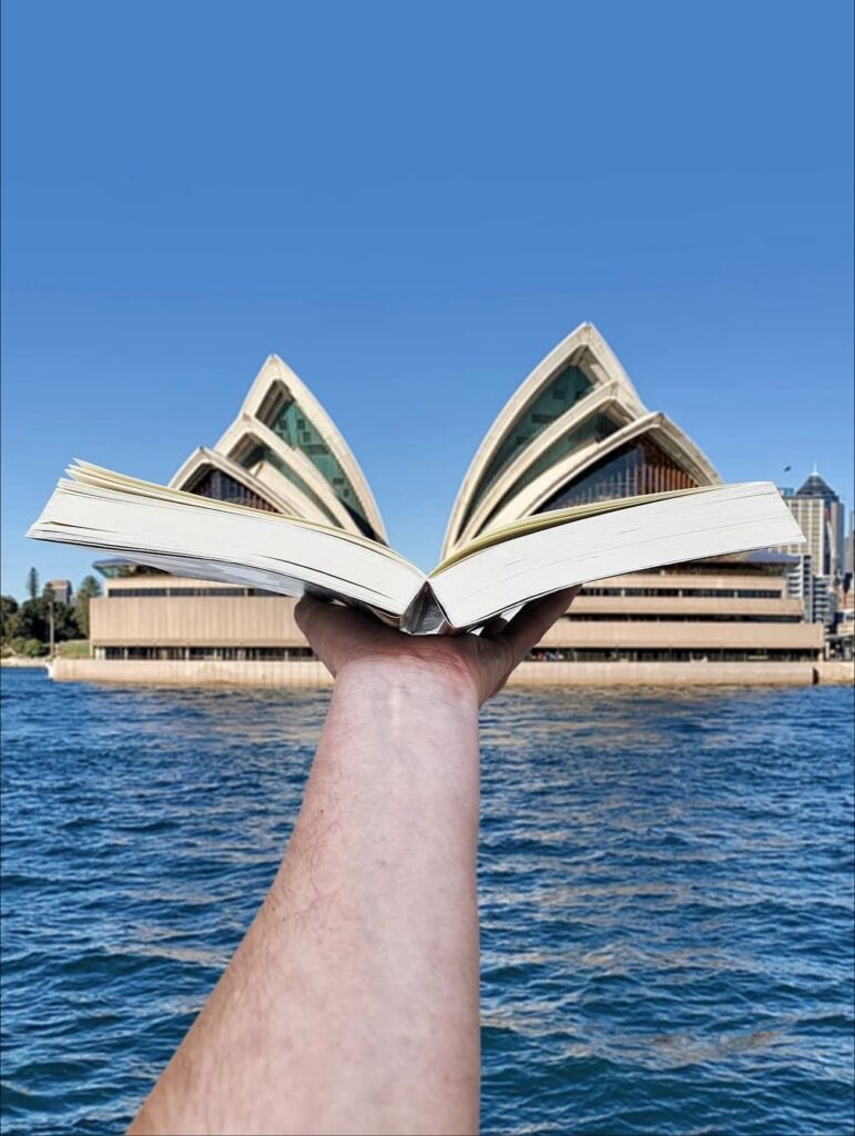 A hand holds an open book at arm’s length so the pages align with the sails of the Sydney Opera House, turning the landmark into a story mid-flight. Architecture becomes literature, rising from the harbor like a narrative unfolding.