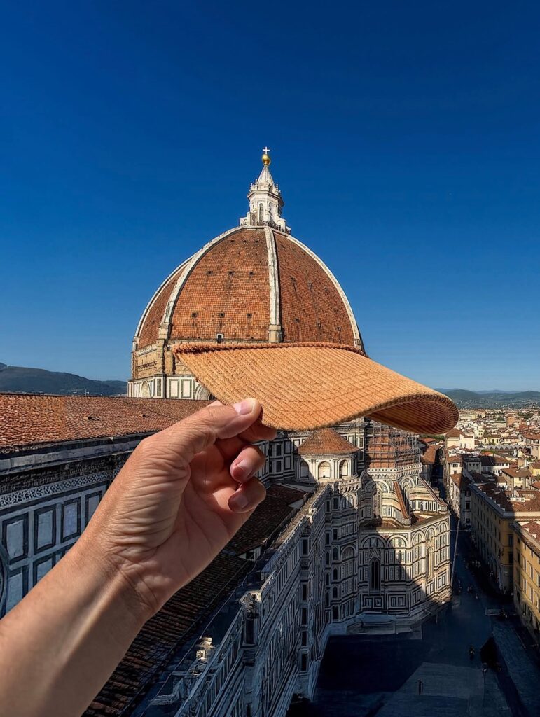 A woven visor is held in front of Florence’s Duomo, matching the dome’s terracotta color and curve so perfectly it feels like the cathedral is casually wearing a sun hat. Renaissance gravity meets vacation whimsy.