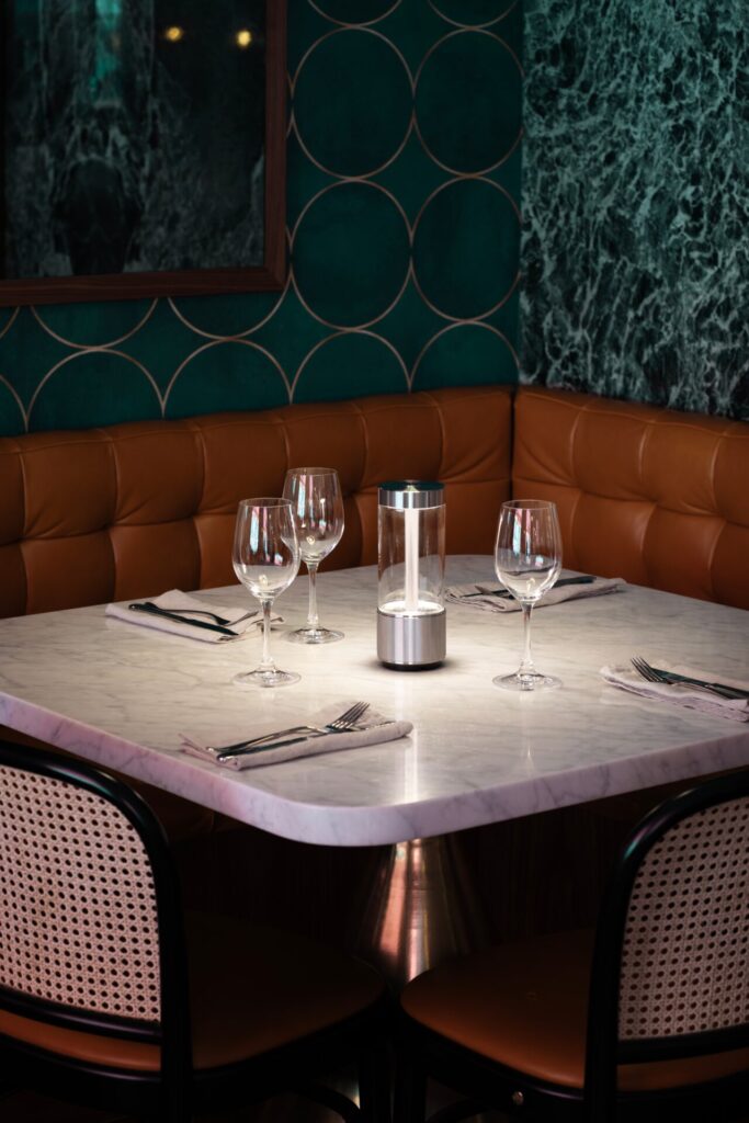 A portable table lamp centered on a marble restaurant table casts a soft, intimate glow over place settings, supporting layered lighting in a hospitality dining environment.