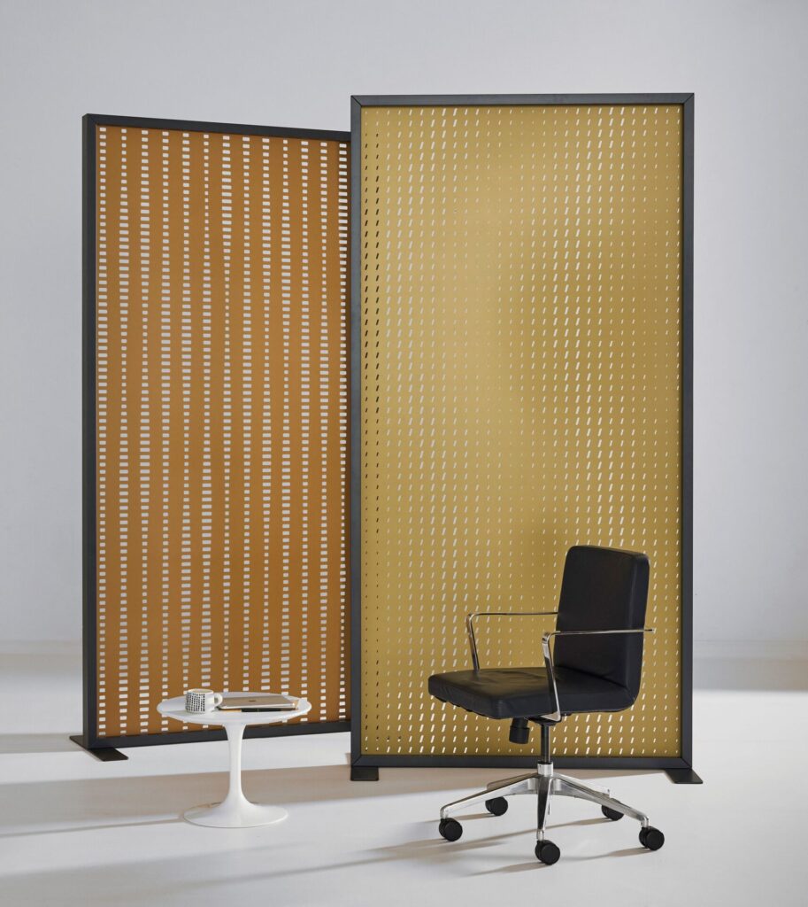 Two warm-toned perforated panels form a gentle backdrop for a compact workspace, their diagonal cutouts catching light like falling dashes. A black task chair and small tulip side table anchor the foreground, underscoring how the screens can carve out privacy without closing a room off.