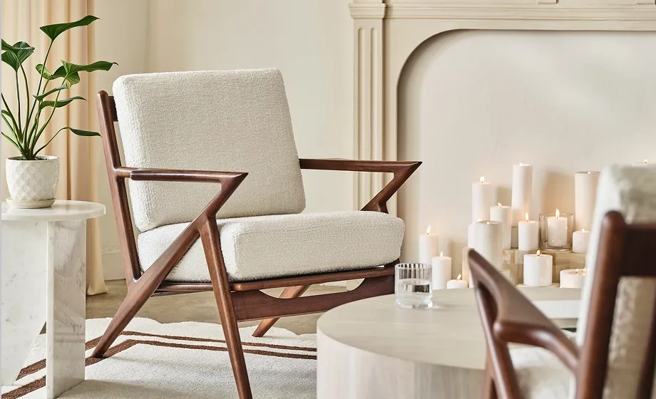 A mid-century–inspired lounge chair pairs warm sculpted wood with Cloud Dancer upholstery, set beside a fireplace lined with candles. The contrast between crisp white texture and rich timber adds calm precision to the room.