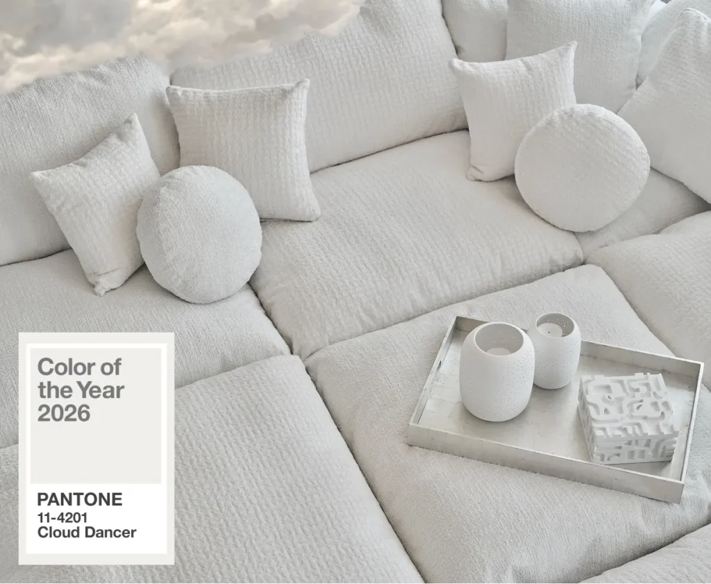 A plush white sectional in Pantone Cloud Dancer fills the frame, layered with tonal pillows and a silver tray holding sculptural ceramics. The textures shift softly across the near-white palette, turning minimalism into a tactile, inviting surface.
