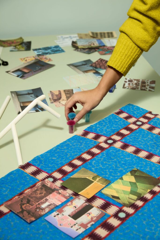 A hand places color poms over a bright Tressage sample while inspiration photos scatter across the table. The crisp burgundy-and-cream grid cuts through the electric blue field, showing how memory, mapping, and material converge in Ritmi’s design process.