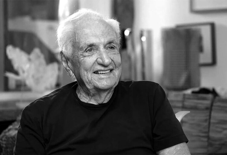 A black-and-white portrait of an older man seated and smiling warmly, captured in a soft, candid moment. The blurred studio background hints at creative tools and materials, giving the scene an intimate, reflective quality.