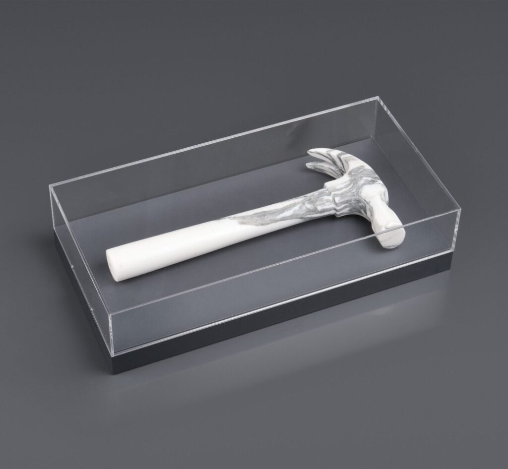 Claw hammer carved from Arabescato Carrara marble, shown in a transparent case that emphasizes precision and material weight.