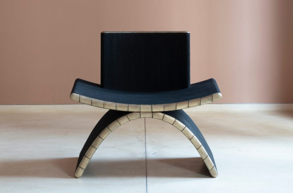 Front view of sculptural lounge seating in the Tarang Chair, highlighting compact proportions, darkened teak grain, and restrained bronze accents..