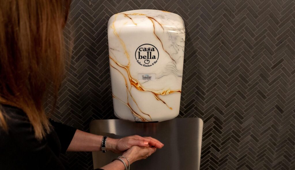 ThinAir hand dryer with custom marble-look cover in use above a stainless splash guard, demonstrating touchless hygiene and design-forward customization in a commercial restroom setting.