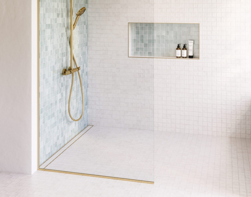 Satin Brass linear shower drain set into pale mosaic tile, softening the technical edge of architectural drainage with a warm metallic finish.