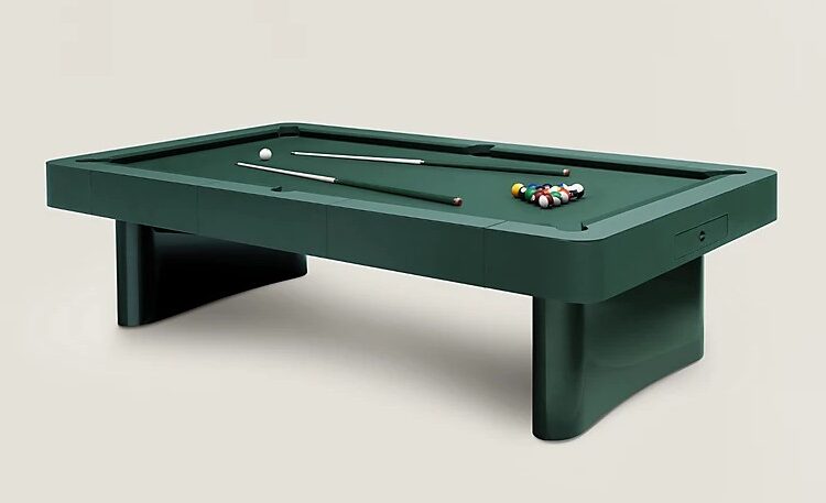 Angled view of Hermès Off Piste pool table with cues and racked balls, showcasing matte leather playing surface and softened corners — designer billiards table